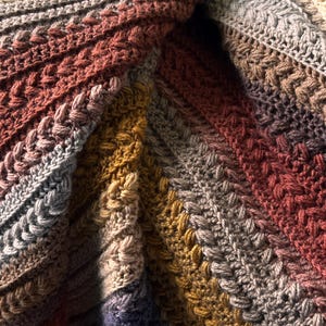 Pode incluir: Close-up de um cobertor de crochê feito à mão com um padrão diagonal texturizado. O cobertor apresenta um gradiente de cores, passando do bege claro a tons de cinza, ferrugem e amarelo mostarda. Os pontos criam um efeito trançado em relevo.