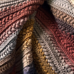 Può includere: Primo piano di una coperta all'uncinetto fatta a mano con un motivo diagonale strutturato. La coperta presenta una miscela di colori caldi, tra cui sfumature di marrone, ruggine, oro e grigio. L'intricato lavoro a punto crea un aspetto accogliente e invitante.