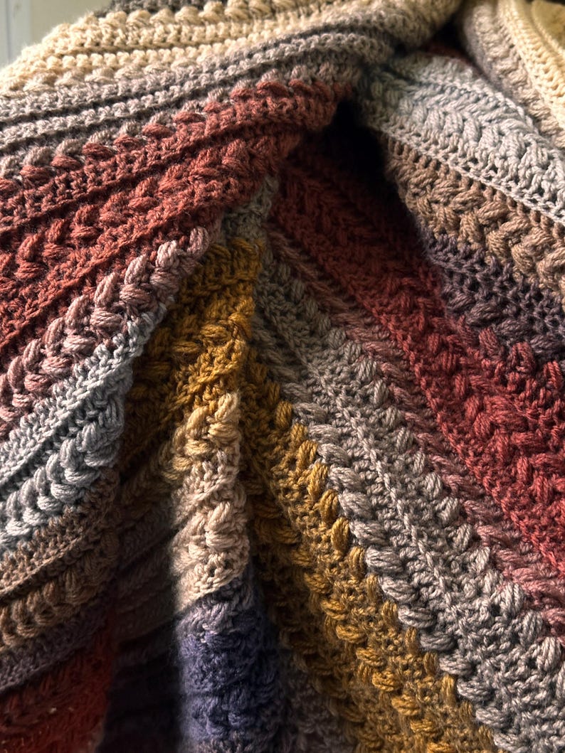 May include: Close-up of a handmade crochet blanket with a textured stitch pattern. The blanket features a gradient of colors, including shades of brown, gray, and mustard yellow. The diagonal design creates a cozy and warm aesthetic.