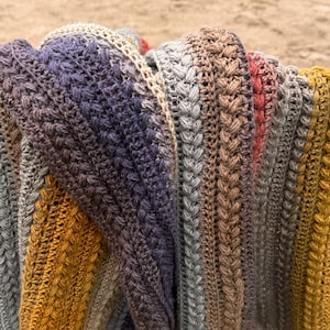 May include: A close-up of a handmade, multi-colored crochet blanket. The blanket features a textured stitch pattern and a gradient of colors, including gray, purple, gold, and red. The blanket is folded, showcasing the intricate design and craftsmanship.