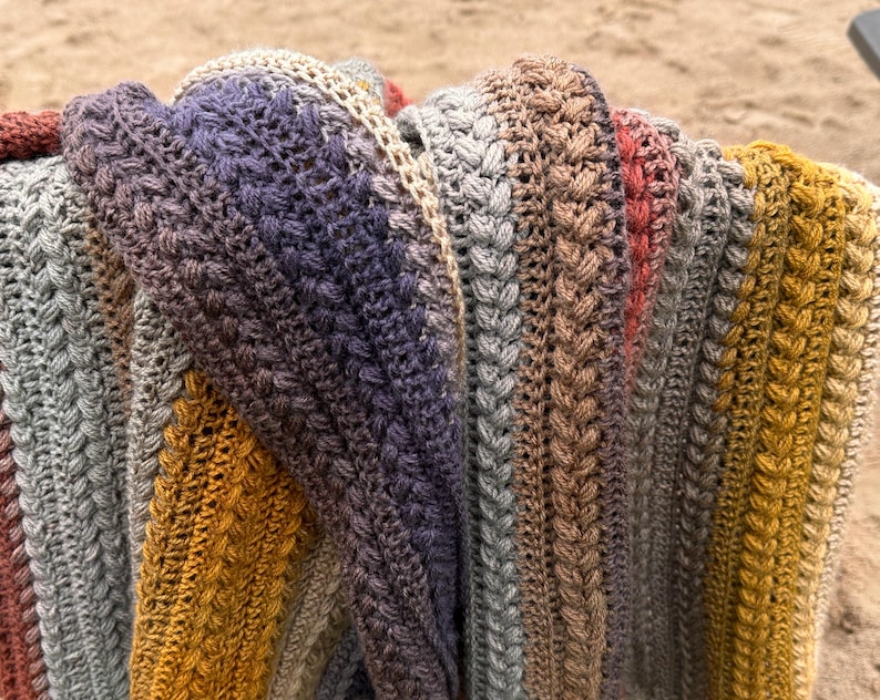 May include: A close-up of a handmade, multi-colored crochet blanket. The blanket features a textured stitch pattern and a gradient of colors, including gray, purple, gold, and red. The blanket is folded, showcasing the intricate design and craftsmanship.