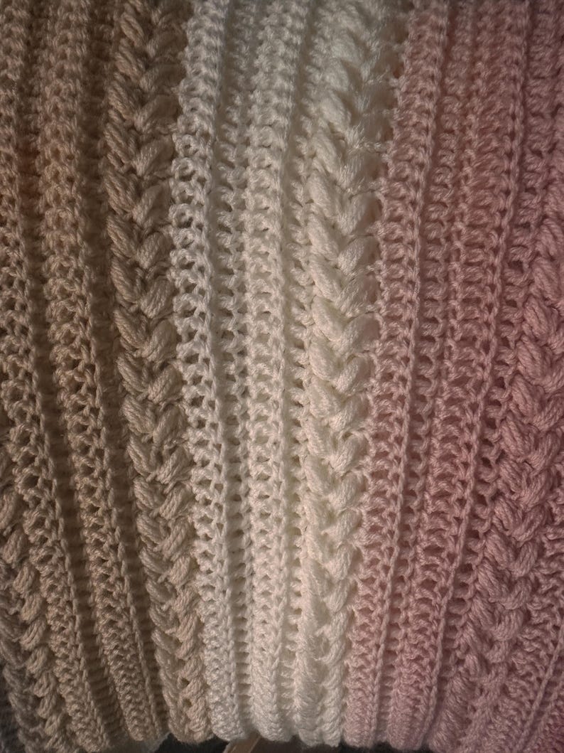K&ouml;nnte beinhalten: Nahaufnahme einer handgefertigten H&auml;keldecke mit Farbverlauf. Die Decke geht von hellem Beige &uuml;ber Wei&szlig; zu einem zarten Rosa &uuml;ber. Das Design zeigt strukturierte vertikale Streifen mit komplizierten Stichmustern.