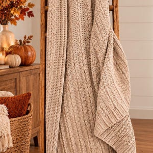 May include: A beige, chunky knit throw blanket featuring a textured cable stitch pattern. The blanket is draped over a tall, vertical wooden ladder, showcasing its thick, soft yarn construction and consistent neutral color throughout the fabric.