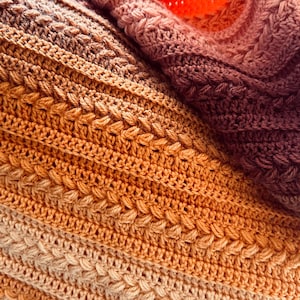 May include: A close-up shot of a handmade crochet blanket in a gradient of warm colours. The blanket transitions from light beige to shades of orange, brown, and deep purple. The textured stitch pattern adds visual interest and depth to the cosy textile.