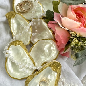 May include: Several oyster shell jewelry dishes with gold rims and pearlescent interiors are arranged on a white cloth. A gold ring and hoop earrings rest in the shells, with a strand of pearls draped across them. Pink and white roses are in the background.