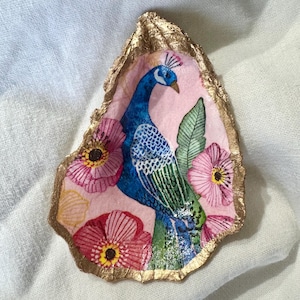 May include: A decorative oyster shell featuring a hand-painted peacock with blue and green plumage, surrounded by pink flowers and green leaves. The shell has a gold-colored edge and a pink background.