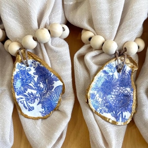 May include: Two napkin rings with a blue and white floral design on an oyster shell, accented with gold trim. The shells are attached to a string of wooden beads, and are wrapped around beige linen napkins.