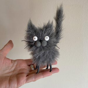 May include: A gray, fuzzy, handmade cat toy with large white eyes and a black nose. The cat toy has black wire whiskers and black legs.