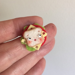 May include: A felt sandwich with a smiling face, cheese, tomato, and lettuce. The sandwich is a cute, handmade craft.