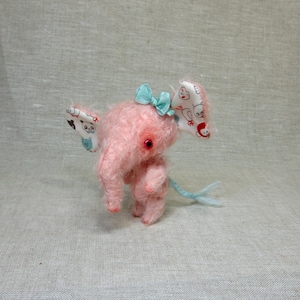 May include: A pink plush elephant toy with a blue bow and fabric ears with a white and blue pattern. The elephant has a long tail with a blue tip.