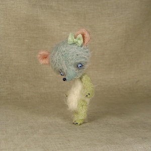 Puede incluir: Un peque&ntilde;o oso de peluche hecho a mano con un cuerpo azul claro y verde, pelaje blanco y un lazo verde claro. El oso tiene ojos azules y una nariz negra.