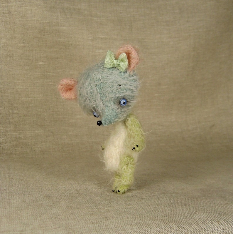 Puede incluir: Un peque&ntilde;o oso de peluche hecho a mano con un cuerpo azul claro y verde, pelaje blanco y un lazo verde claro. El oso tiene ojos azules y una nariz negra.