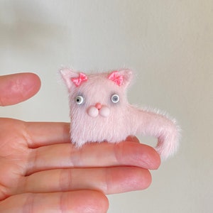 May include: A pink fuzzy cat toy with white eyes and pink bows on its ears. The toy is sitting on a person's hand.