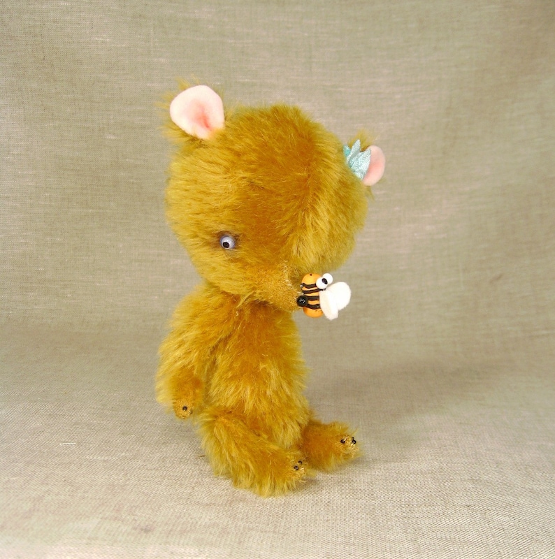 Puede incluir: Un peque&ntilde;o osito de peluche marr&oacute;n dorado con una flor azul claro en la cabeza. El oso tiene un amuleto de abeja en la nariz.