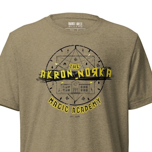 Könnte beinhalten: Ein khaki-grünes T-Shirt mit einem gelben Grafikdesign eines Schulgebäudes mit dem Text "The Akron Nozka Magic Academy Est. 1900" in einem Kreis.