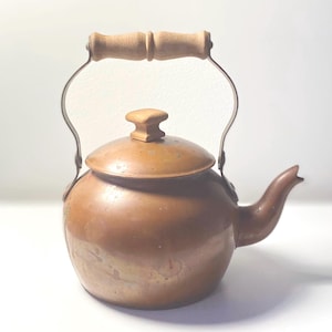 May include: A copper tea kettle with a rounded body, a curved spout, and a removable lid featuring a light-colored wooden knob. The kettle includes a metal handle with a central light-colored wooden grip for lifting and pouring.