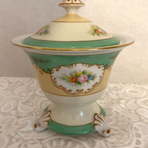 May include: A vintage porcelain sugar bowl with a lid. The bowl is decorated with floral patterns in shades of pink, yellow, and green. The bowl has a green base with gold accents and three feet.