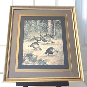 May include: A framed print featuring a group of wild turkeys in a forest setting. The artwork displays several turkeys in shades of brown, black, and white, surrounded by trees and foliage. The print is encased in a gold-toned frame with a multi-layered matting in dark gray and gold.