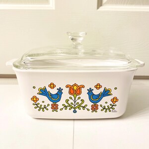May include: A white rectangular glass casserole dish with a clear glass lid. The front features a colorful graphic print of two blue birds facing a central orange and yellow tulip, surrounded by small green leaves and orange flowers. Text reads 1973 CORNING.