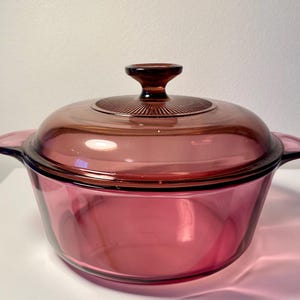 May include: A vintage, ruby-colored glass casserole dish with a matching lid. The dish has two handles and a round shape. The lid features a dark brown knob and a ridged design. This cookware is ideal for baking and serving.