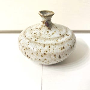 May include: A small, round ceramic vase with a speckled off-white glaze and brown spots. The vase features a narrow neck with a flared rim and a thin wire wrapped around the neck, adorned with a single, small, smooth, dark brown oval bead.