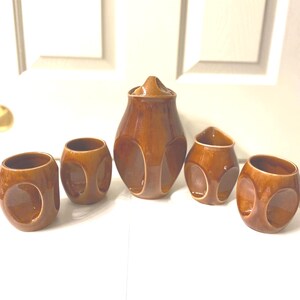 May include: A ceramic beverage set featuring a tall lidded pitcher, a smaller creamer pitcher, and three matching cups. Each piece is finished in a glossy, warm brown glaze and features a distinct, recessed circular cutout design on the sides.