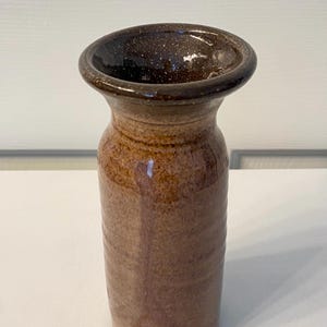 May include: A cylindrical ceramic vase with a speckled dark brown glazed rim and a textured, earthy tan body. The vase features a wide, flared opening that tapers into a straight, vertical neck and a slightly wider base, showcasing a rustic finish.
