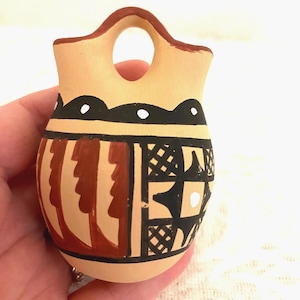 May include: A small, tan ceramic vessel with a single arched handle at the top. The body features hand-painted geometric patterns in black and reddish-brown, including scalloped lines with white dots and a grid-like design on the lower section.