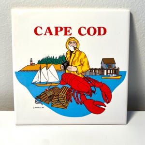 May include: Square ceramic tile with a colorful illustration of Cape Cod. The image features a fisherman in a yellow coat, a red lobster, a lighthouse, boats, and the words "CAPE COD" in red.