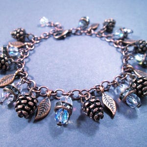 May include: A copper-toned charm bracelet featuring pinecone and leaf charms, interspersed with clear and blue crystal beads. The bracelet is approximately 7 inches in length, with a lobster claw clasp.