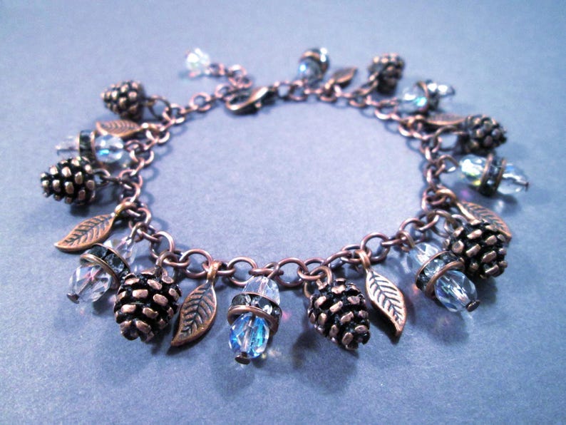 May include: A copper-toned charm bracelet featuring pinecone and leaf charms, interspersed with clear and blue crystal beads. The bracelet is approximately 7 inches in length, with a lobster claw clasp.