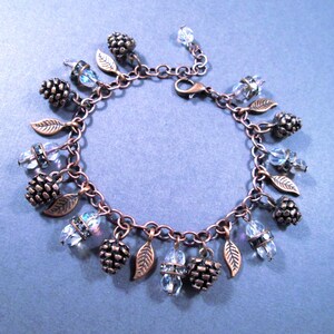 May include: A copper-toned charm bracelet featuring a series of decorative elements. The bracelet includes small pinecone charms, leaf charms, and clear, faceted crystal beads. The bracelet is displayed on a blue surface.
