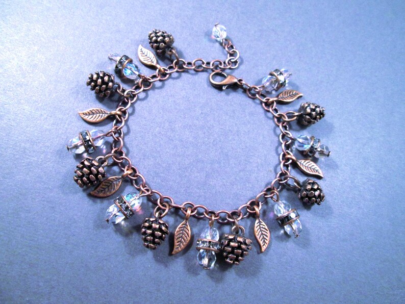 May include: A copper-toned charm bracelet featuring a series of decorative elements. The bracelet includes small pinecone charms, leaf charms, and clear, faceted crystal beads. The bracelet is displayed on a blue surface.