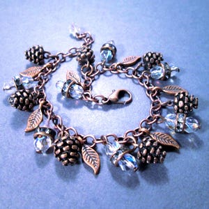 May include: A copper-toned charm bracelet featuring pinecone and leaf charms, along with clear, faceted crystal beads. The bracelet has a lobster clasp closure and is displayed on a blue-gray surface. The bracelet is a nature-inspired jewelry piece.
