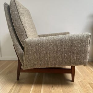 May include: A mid-century modern armchair with a light brown tweed fabric and dark wooden legs. The chair has a high back and armrests, offering a comfortable seating experience. The design is simple yet elegant, suitable for a living room or lounge area.
