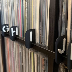 Puede incluir: Separadores de discos de vinilo negros con letras blancas de la F a la K se muestran frente a una colección de discos de vinilo. Los separadores están en un estante, lo que ayuda a organizar los discos alfabéticamente.
