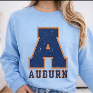 May include: Pale blue sweatshirt featuring a large navy blue letter "A" and the word "AUBURN" in a distressed, collegiate style. The "A" has an orange outline. The sweatshirt is a crew neck style.