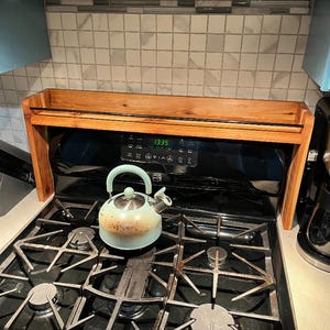 Puede incluir: Un estante de madera para la estufa con una tetera turquesa en la parte superior. El estante está instalado sobre una estufa de gas negra con cuatro quemadores. La estufa tiene una pantalla digital que muestra "1:35".