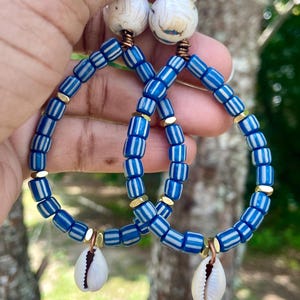 May include: A pair of hoop earrings featuring blue and white striped glass beads, small metallic gold spacers, a large white marbled bead at the top, and a single white cowrie shell dangling from the bottom of each hoop, secured with copper wire.