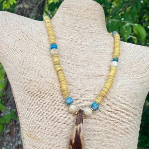 May include: A handcrafted necklace featuring a pendant and a strand of yellow, round beads. The necklace includes blue and white accent beads. The pendant is brown with a unique shape. The necklace is displayed on a mannequin.