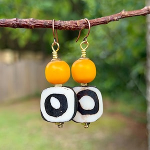 May include: A pair of dangle earrings featuring a bright yellow spherical bead positioned above a flat, square-shaped white bead with a prominent black circular pattern in the center, all connected by gold-toned metal findings and copper-toned hooks.