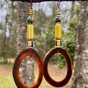 May include: A pair of brown oval hoop earrings with gold accents. The earrings feature a yellow, white, blue, and green beaded dangle.