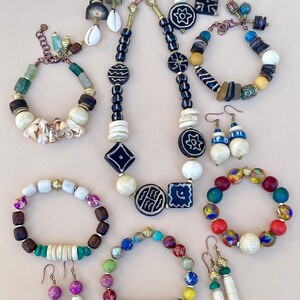 May include: A collection of handcrafted jewelry, including necklaces, bracelets, and earrings. The pieces feature a variety of beads in white, black, blue, green, and red, with accents of gold and shell. The designs showcase geometric and organic shapes.