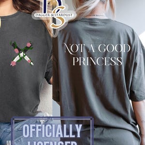 May include: Dark grey t-shirt featuring an embroidered design of crossed daggers and flowers. The back of the shirt displays the text "NOT A GOOD PRINCESS". The shirt also has the text "COMFORT COLORS" and "OFFICIALLY LICENSED".
