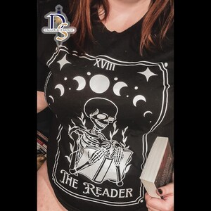 K&ouml;nnte beinhalten: Schwarzes T-Shirt mit einem wei&szlig;en Grafikdruck eines Skeletts, das ein Buch liest. Die Grafik enth&auml;lt den Text "The Reader" und "XVIII".