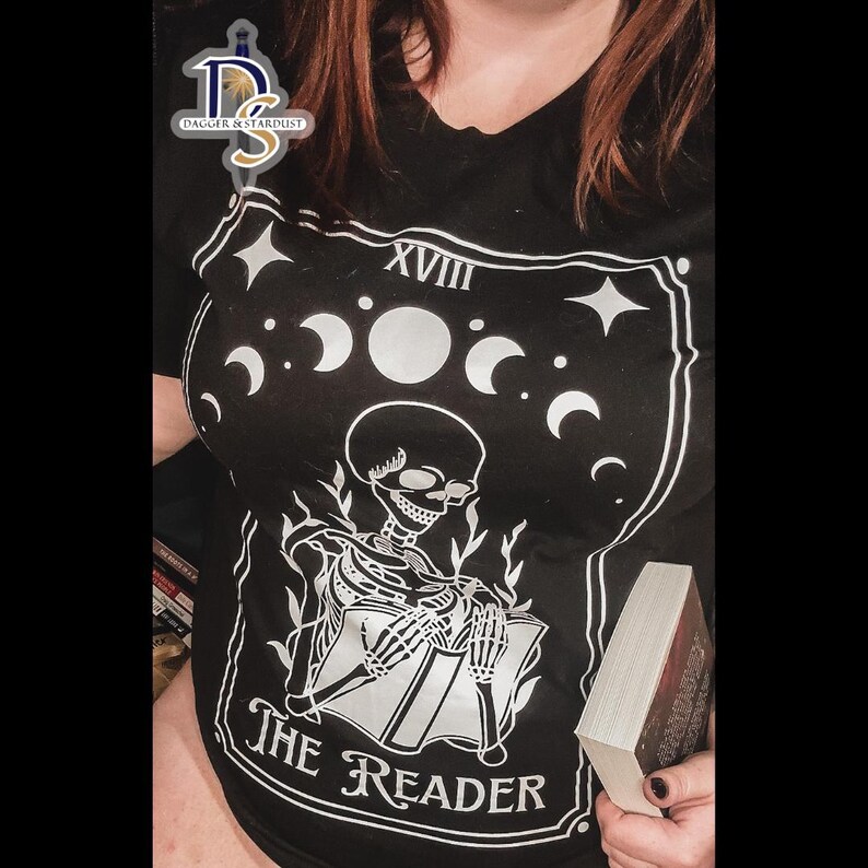 K&ouml;nnte beinhalten: Schwarzes T-Shirt mit einem wei&szlig;en Grafikdruck eines Skeletts, das ein Buch liest. Die Grafik enth&auml;lt den Text "The Reader" und "XVIII".