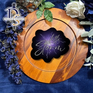 May include: A black and purple round sticker with the text "little storm" in silver script. The sticker has a lightning bolt design.