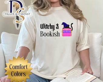 May include: Cream-colored t-shirt with the text "Witchy & Bookish" and a graphic of a black cat wearing a witch hat on top of stacked books. The shirt also features the words "Comfort Colors" in a gold starburst.