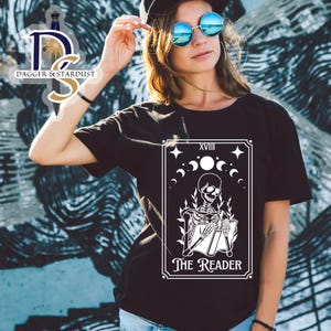 K&ouml;nnte beinhalten: Schwarzes T-Shirt mit einem wei&szlig;en Grafikdruck einer Tarotkarte. Die Karte ist "Der Leser", die 18. Karte der Gro&szlig;en Arkana. Die Karte zeigt ein Skelett, das ein Buch liest.