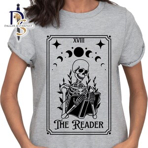 K&ouml;nnte beinhalten: Ein graues T-Shirt mit einer schwarz-wei&szlig;en Illustration einer Tarotkarte. Die Karte ist mit "XVIII The Reader" beschriftet und zeigt ein Skelett, das ein Buch h&auml;lt.